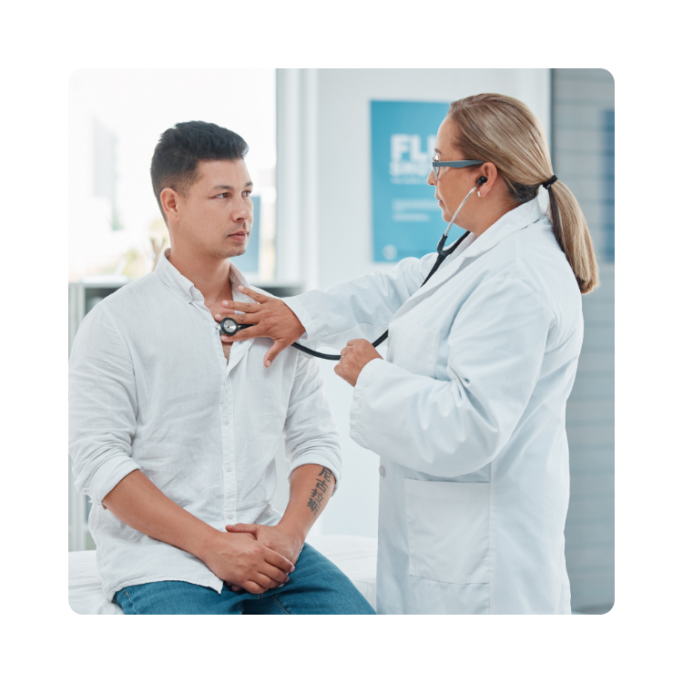 Lekarka w białym fartuchu i maseczce osłuchuje stetoskopem klatkę piersiową siedzącego pacjenta podczas badania w poradni medycyny pracy