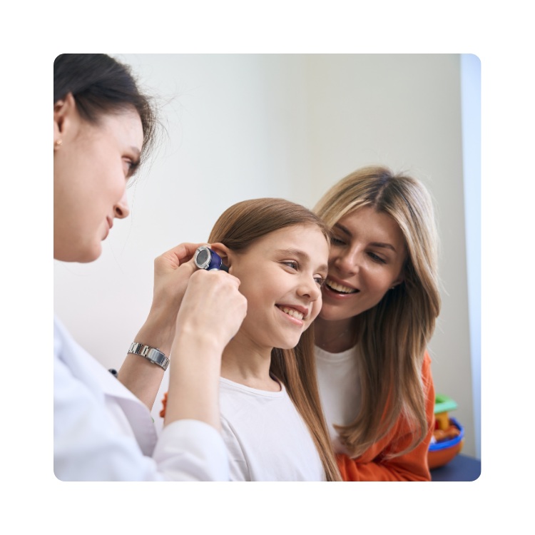 Lekarka bada ucho uśmiechniętej dziewczynki otoskopem, obok stoi mama wspierająca c&oacute;rkę podczas wizyty w poradni laryngologicznej.